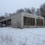 Zema viena stāva betona ēka ar metāla restotiem logiem un grafiti uz fasādes Mazās Stacijas ielas apkaimē Rīgā. Apkārt redzams sniegs, koki un daudzdzīvokļu ēkas fonā, kā arī stāvēšanai novietots automobilis