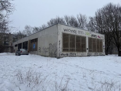 Zema viena stāva betona ēka ar metāla restotiem logiem un grafiti uz fasādes Mazās Stacijas ielas apkaimē Rīgā. Apkārt redzams sniegs, koki un daudzdzīvokļu ēkas fonā, kā arī stāvēšanai novietots automobilis