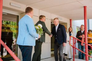 Cilvēku grupa stāv uz ēkas ieejas kāpnēm, redzamas sarkanas margas, stikla durvis un gaiša fasāde, viens cilvēks tur rokās baltu ziedu pušķi
