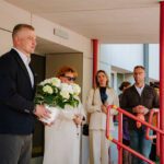 Cilvēku grupa stāv pie ēkas ieejas ar sarkanām margām; priekšplānā viens cilvēks tur baltu ziedu pušķi, kamēr citi stāv rindā gar margām; fonā redzama gaiša ēkas fasāde un stikla durvis.