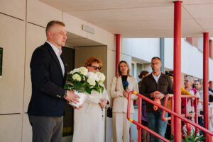 Cilvēku grupa stāv pie ēkas ieejas ar sarkanām margām; priekšplānā viens cilvēks tur baltu ziedu pušķi, kamēr citi stāv rindā gar margām; fonā redzama gaiša ēkas fasāde un stikla durvis.