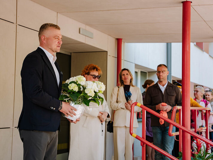 Cilvēku grupa stāv pie ēkas ieejas ar sarkanām margām; priekšplānā viens cilvēks tur baltu ziedu pušķi, kamēr citi stāv rindā gar margām; fonā redzama gaiša ēkas fasāde un stikla durvis.