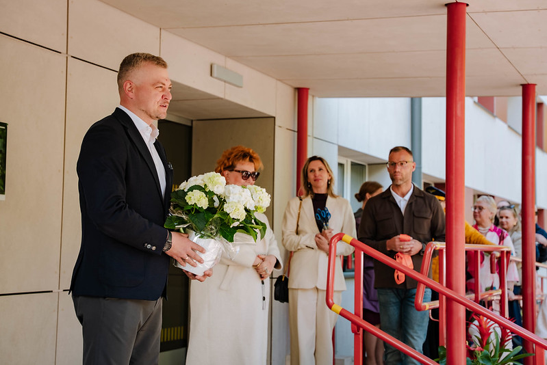 Cilvēku grupa stāv pie ēkas ieejas ar sarkanām margām; priekšplānā viens cilvēks tur baltu ziedu pušķi, kamēr citi stāv rindā gar margām; fonā redzama gaiša ēkas fasāde un stikla durvis.