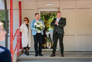 Trīs cilvēki stāv uz ēkas ieejas kāpnēm pie stikla durvīm; viens tur rokās baltu ziedu pušķi, blakus redzamas sarkanas margas un gaiša ēkas fasāde
