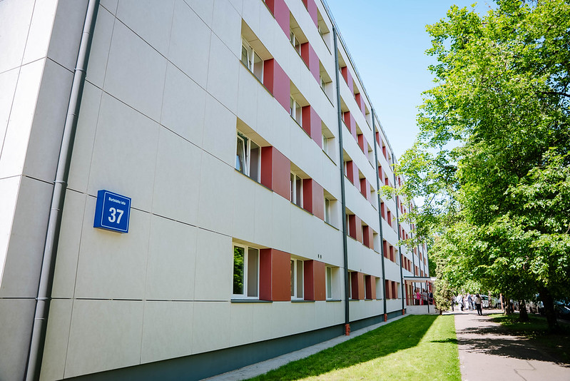 Daudzstāvu ēka ar baltu un sarkanīgu fasādi un logu rindām; uz sienas redzama zilā ielas numura zīme “37”; gar ēku ved zaļš zāliens un gājēju celiņš ar kokiem, tālumā redzami cilvēki pie ieejas