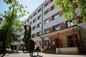 Daudzstāvu ēka ar baltu un sarkanīgu fasādi, pie kuras ieejas pulcējušies vairāki cilvēki; ēku ieskauj koki un saulaina vide