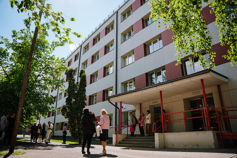 Daudzstāvu ēka ar baltu un sarkanīgu fasādi, pie kuras ieejas pulcējušies vairāki cilvēki; ēku ieskauj koki un saulaina vide