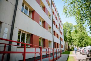 Daudzstāvu ēkas fasāde baltā un sarkanīgā krāsā ar logu rindām; gar ēku ved ietve un piebraucamais ceļš, redzama sarkana pandusa ar margām, koki un daži novietoti automobiļi
