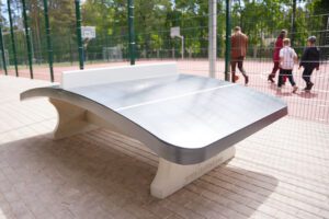 Āra galda tenisa galds pie sporta laukuma ar nožogotu basketbola laukumu fonā.