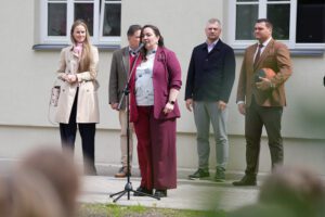 Persona runā pie mikrofona pie ēkas, fonā stāv vairāki cilvēki; viens tur basketbola bumbu.