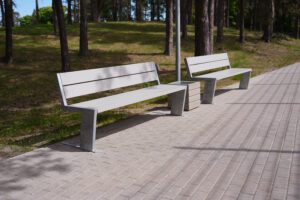 Soliņi un atkritumu tvertne gar bruģētu celiņu parkam līdzīgā teritorijā ar kokiem fonā