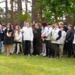 Skolēni rindā skolas pagalmā, vairāki tur rokās basketbola bumbas, fonā koki.