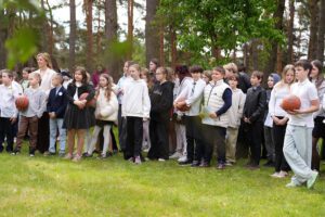 Skolēni rindā skolas pagalmā, vairāki tur rokās basketbola bumbas, fonā koki.