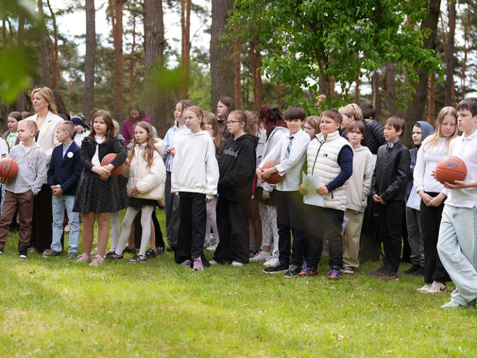 Skolēni rindā skolas pagalmā, vairāki tur rokās basketbola bumbas, fonā koki.