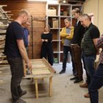Cilvēku grupa darbnīcā stāv ap darba galdu un klausās skaidrojumu par restaurējamu loga rāmi, fonā plaukti ar materiāliem.