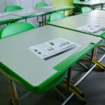 Tukša klases telpa ar zaļiem skolēnu galdiem un krēsliem; uz galdiem izvietoti drukāti materiāli