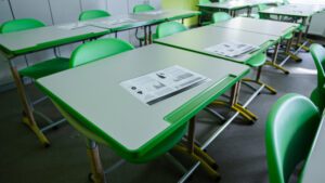 Tukša klases telpa ar zaļiem skolēnu galdiem un krēsliem; uz galdiem izvietoti drukāti materiāli