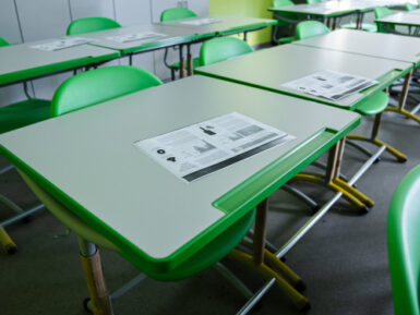 Tukša klases telpa ar zaļiem skolēnu galdiem un krēsliem; uz galdiem izvietoti drukāti materiāli