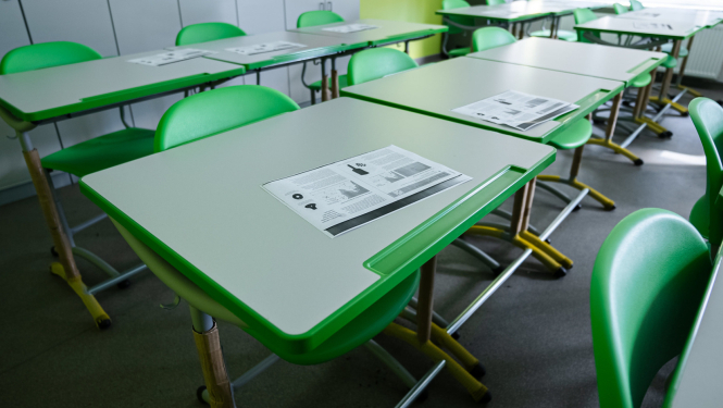 Tukša klases telpa ar zaļiem skolēnu galdiem un krēsliem; uz galdiem izvietoti drukāti materiāli