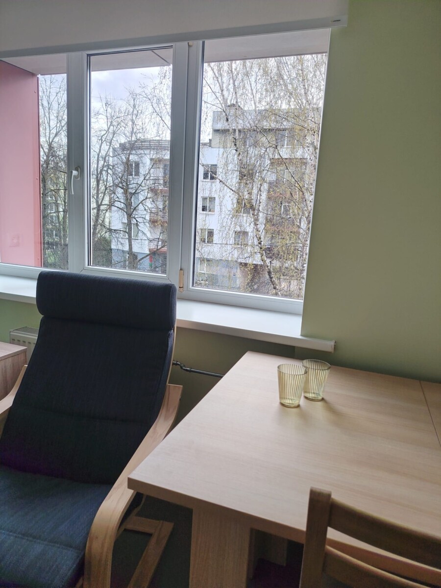 Kabinets ar galdu, krēslu un logu, aiz kura redzami koki un dzīvojamā ēka
