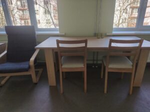 Galds ar trim krēsliem pie logiem, aiz kuriem redzami koki un dzīvojamā ēka