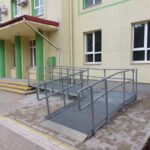 Ēkas ieeja ar metāla pandusu, margām un marķētu piekļuves ceļu.