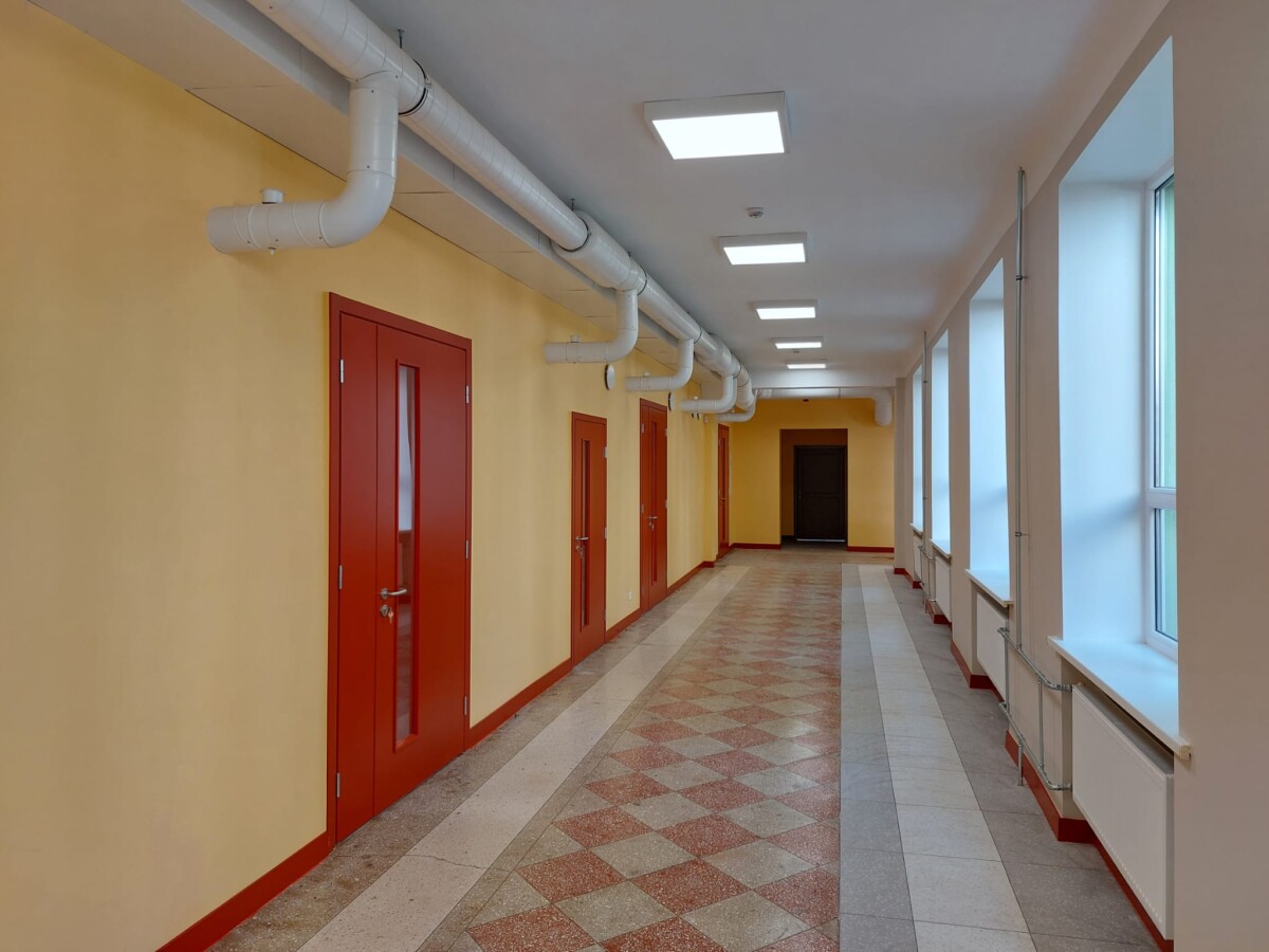 Gaišs gaitenis ar sarkanām durvīm, logiem, radiatoriem un ventilācijas caurulēm pie griestiem.