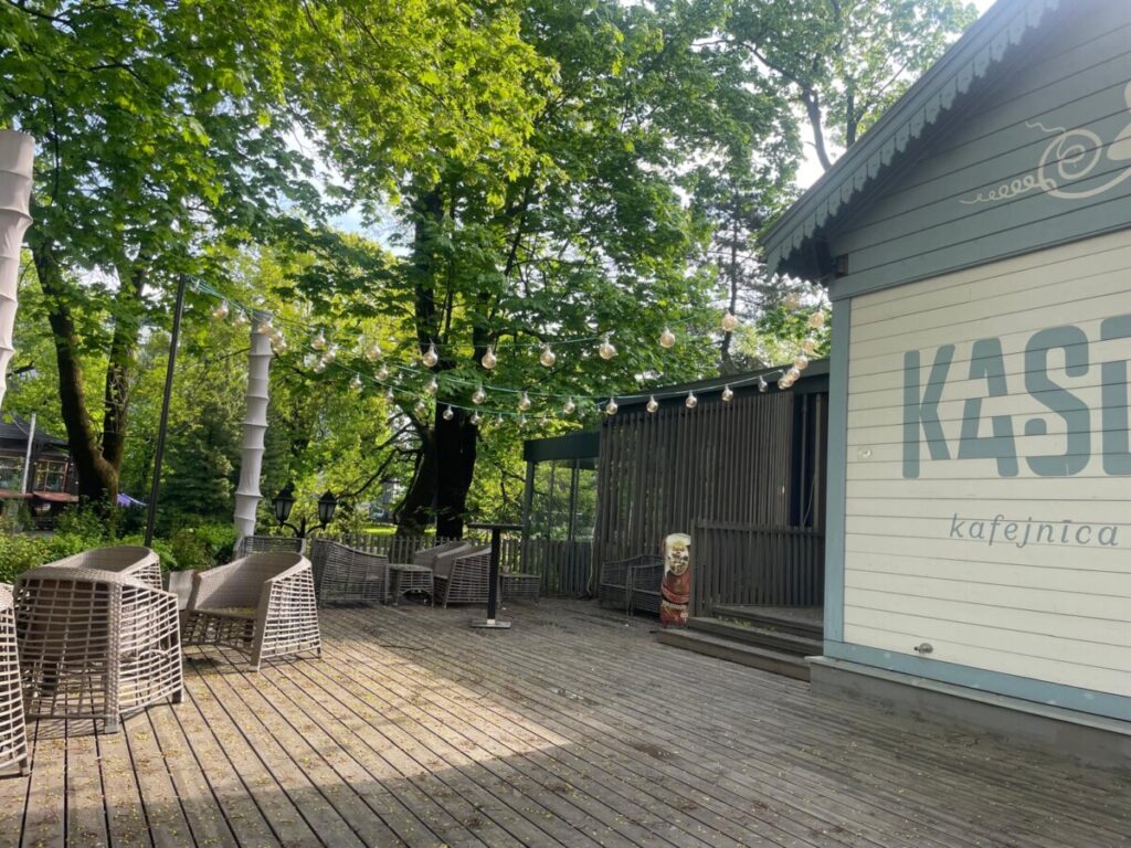 Koka terase pie kafejnīcas parkā ar pītiem krēsliem un galdiem. Virs terases starp lieliem, zaļiem kokiem izkārti dekoratīvi gaismas virteņu lukturi. Attēla labajā pusē redzama gaiša kafejnīcas ēka ar uzrakstu “KASE kafejnīca”. Saulaina diena, terasi ieskauj zaļumi un koku ēnas.