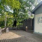 Koka terase pie kafejnīcas parkā ar pītiem krēsliem un galdiem. Virs terases starp lieliem, zaļiem kokiem izkārti dekoratīvi gaismas virteņu lukturi. Attēla labajā pusē redzama gaiša kafejnīcas ēka ar uzrakstu “KASE kafejnīca”. Saulaina diena, terasi ieskauj zaļumi un koku ēnas.