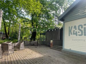 Koka terase pie kafejnīcas parkā ar pītiem krēsliem un galdiem. Virs terases starp lieliem, zaļiem kokiem izkārti dekoratīvi gaismas virteņu lukturi. Attēla labajā pusē redzama gaiša kafejnīcas ēka ar uzrakstu “KASE kafejnīca”. Saulaina diena, terasi ieskauj zaļumi un koku ēnas.