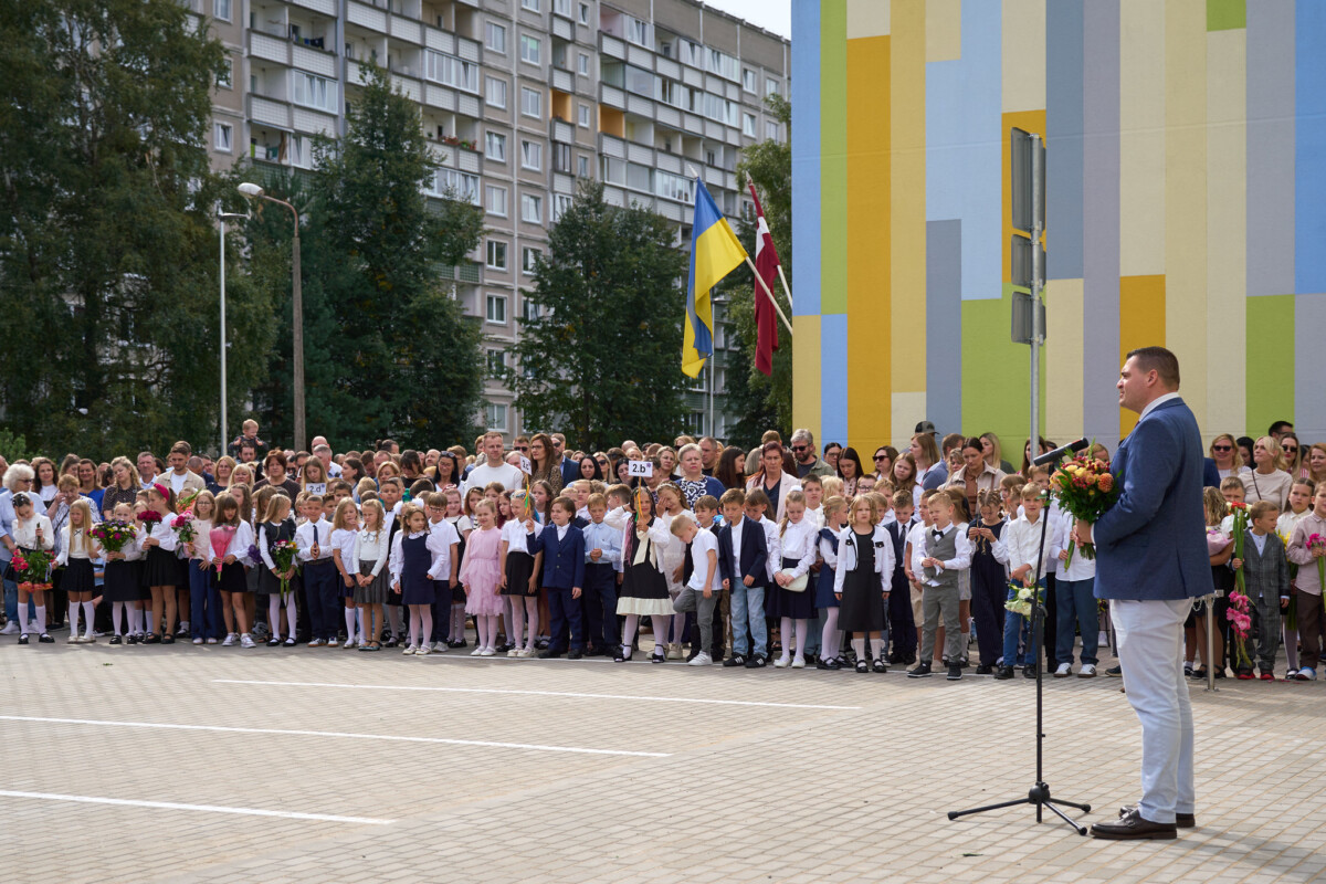 Pie skolas ēkas uzrunu pie mikrofona saka cilvēks ar ziedu pušķi rokās, kamēr pagalmā pulcējušies skolēni, vecāki un pedagogi