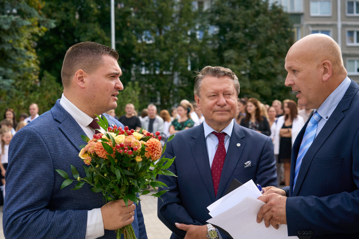Trīs cilvēki sarunājas ārā pie skolas. Viens tur rokās ziedu pušķi, bet otrs dokumentus. Fonā redzami pasākuma apmeklētāji un koki.