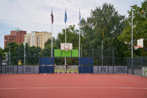 Skolas sporta laukumā redzams basketbola grozs ar vairākiem basketbola bumbu komplektiem pie nožogotā laukuma ieejas