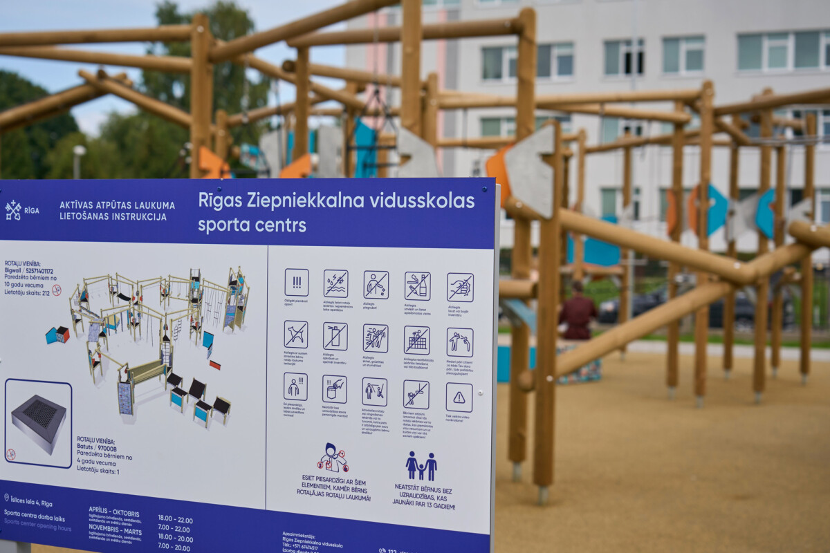 Informatīvs stends ar Rīgas Ziepniekkalna vidusskolas sporta centra lietošanas noteikumiem pie aktīvās atpūtas laukuma. Fonā redzama koka rotaļu un vingrošanas konstrukcija
