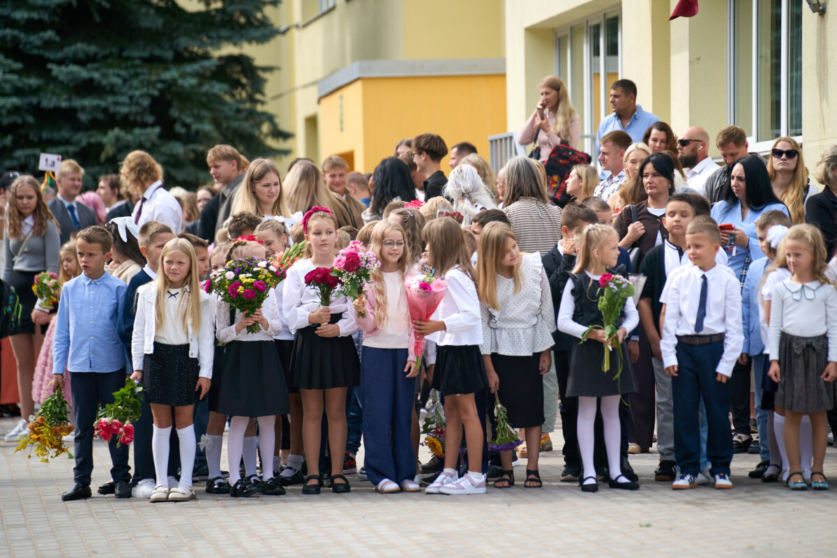 Skolas pagalmā pulcējušies skolēni ar ziedu pušķiem, bet aiz viņiem stāv vecāki un citi pasākuma apmeklētāji pie skolas ēkas
