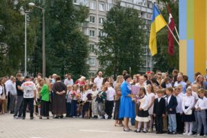 Skolas pagalmā pulcējušies skolēni, pedagogi un ģimenes locekļi svinīgā pasākumā pie Rīgas Ziepniekkalna vidusskolas ēkas. Priekšplānā redzami bērni svētku apģērbā un ar ziediem, fonā plīvo Latvijas un Ukrainas karogi